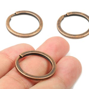 May include: Three bronze-coloured key rings are shown, one held in a hand. The rings are circular with a small opening. They have a rustic, antique appearance. The other two rings are on a white surface. Ideal for keys or small accessories.