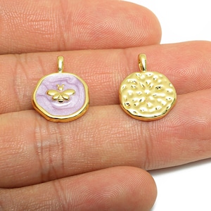 Pode incluir: Dois pingentes dourados. Um é um círculo texturizado. O outro é um pingente dourado, de cor lavanda, com um desenho de abelha dourada. Ambos os pingentes têm uma pequena argola no topo para pendurar.