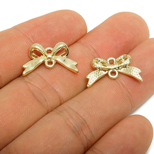 Peut inclure: Deux petits breloques en forme de nœud papillon dorés avec des surfaces texturées. Chaque breloque a une boucle en haut et en bas pour l'attacher à des bijoux ou autres créations. Les nœuds sont tenus dans une main sur fond blanc.