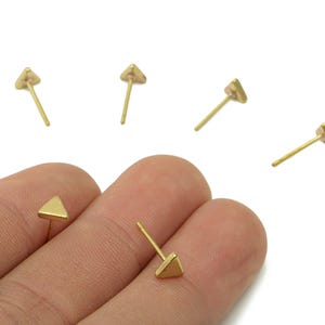 May include: Gold-colored triangle stud earrings. The earrings have a simple geometric design with a small, raised triangle shape on a post. Several earrings are scattered on a white background.