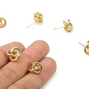 Puede incluir: Pendientes de botón dorados con forma de nudo. Los pendientes tienen un diseño de nudo retorcido y un poste para orejas perforadas. Varios pendientes están esparcidos sobre un fondo blanco, con dos sostenidos en una mano.