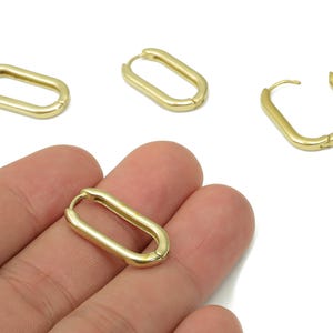 May include: Gold-colored oval hoop earrings. The earrings have a simple, minimalist design with a smooth, polished finish. Several earrings are displayed, with one held in a hand. The earrings are a classic jewelry item.