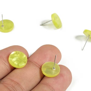 Pode incluir: Uma coleção de brincos de botão amarelo-esverdeados com pinos de prata. Os brincos têm um design redondo e plano com aparência marmorizada. Os brincos são mostrados em um fundo branco.