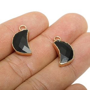 May include: Two crescent moon-shaped pendants. Each pendant features a faceted black gemstone set in a gold-colored metal frame. The pendants have a small loop at the top for attaching to a chain or other jewelry component.