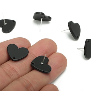 May include: Black heart-shaped stud earrings. The earrings are made of a matte black material and have a silver post. Several earrings are scattered on a white background, with some held in a hand.