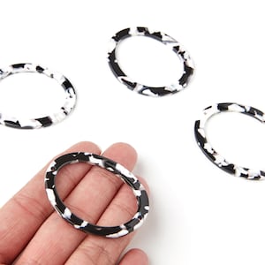 Peut inclure: Quatre composants de bijoux ovales en résine noire et blanche. Les composants présentent un motif moucheté et sont présentés sur un fond blanc. L'un est tenu dans une main.