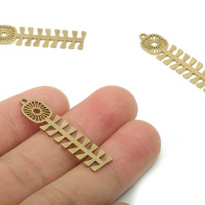 Puede incluir: Tres dijes de metal color latón. Cada dije tiene una parte superior circular con un diseño de rayos de sol y un cuerpo rectangular con una serie de pequeños recortes angulados. Un dije se sostiene en una mano.