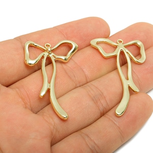 Peut inclure: Deux breloques dorées en forme de nœud papillon. Chaque breloque a une boucle en haut pour être suspendue et un design ouvert et fluide. Les breloques sont affichées sur un fond clair.
