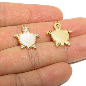 May include: Two gold-colored turtle charms. One charm features a white shell with a gold border and clear crystals. The other charm is solid gold. Both charms have a loop for attaching to jewelry.