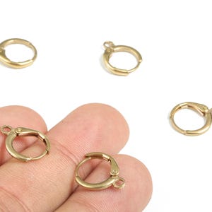 Peut inclure: Plusieurs apprêts de boucles d'oreilles à levier de couleur or sont présentés sur fond blanc. L'un est tenu dans une main. Les apprêts sont petits, circulaires et ont une fermeture à charnière avec une boucle.