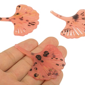 May include: Three translucent, pink ginkgo leaf-shaped pendants with dark brown spots. One pendant is held in a hand, while two others are displayed separately. The pendants have a stem and a fan-like shape with delicate lines.