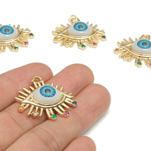 May include: Gold-colored eye-shaped charms with a blue iris and white background. The charms have colorful gemstone accents and are designed with a sunburst pattern. The charms are for jewelry making.
