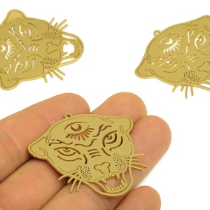 Puede incluir: Tres colgantes de metal dorado con forma de cabeza de pantera estilizada con intrincados detalles recortados. Los colgantes presentan bocas abiertas, dientes visibles y un diseño de tercer ojo. Un colgante se sostiene en una mano.