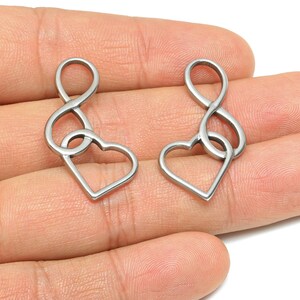 May include: Two silver-colored heart-shaped pendants with an infinity symbol design. The pendants are made of a shiny metal and are displayed against a light background. The jewelry is a romantic accessory.