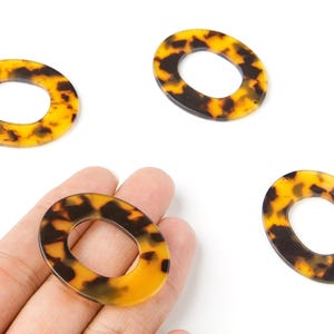 Peut inclure: Quatre composants de bijoux ovales avec un motif écaille de tortue. Les composants sont un mélange de jaune, de marron et de noir. Un composant est tenu dans une main. Ces composants sont probablement utilisés pour la fabrication de bijoux ou l'artisanat.