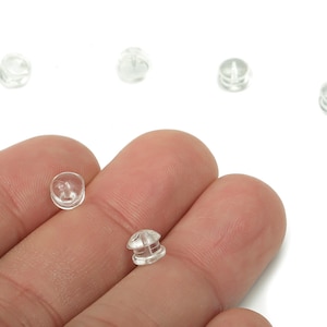 May include: Clear, small earring backs are displayed against a white background. Several are scattered, with two held between fingers. The earring backs are made of a transparent material, and are round with a small post hole.
