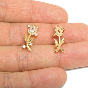May include: Two gold-colored tulip-shaped charms. One charm features a clear, oval-shaped stone surrounded by smaller clear stones. The other charm is a cutout of a tulip. Both charms have a stem and leaves.
