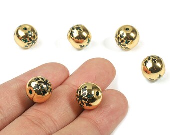 Puede incluir: Cuentas redondas doradas con un diseño floral en negro y verde. Las cuentas se sostienen en una mano, con varias más dispersas sobre una superficie blanca. Las cuentas tienen una superficie brillante y reflectante.