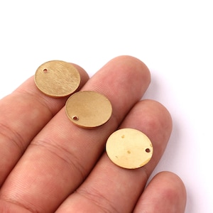 Può includere: Tre piccoli ciondoli rotondi in metallo color oro con finitura spazzolata. Ogni ciondolo ha un piccolo foro vicino al bordo. I ciondoli sono tenuti in mano su sfondo bianco. Questi sono probabilmente materiali per la creazione di gioielli.