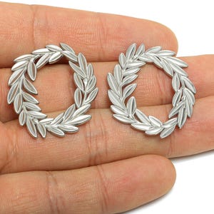Peut inclure: Deux pendentifs argentés en forme de couronne circulaire. Chaque pendentif présente un motif détaillé de feuilles disposées en cercle. Les pendentifs sont présentés sur un fond clair.