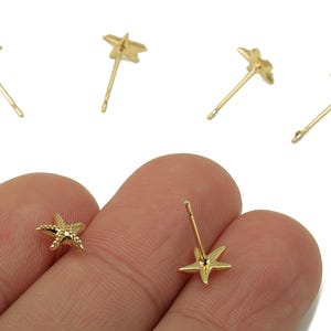 May include: Gold-colored starfish stud earrings. The earrings have a textured surface and a post backing. The image shows several pairs of the earrings, with one pair held between fingers.