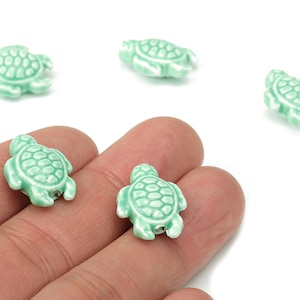 May include: A collection of small, pale green turtle-shaped beads. Each bead features a textured shell pattern and small holes for threading. The beads are shown against a white backdrop.