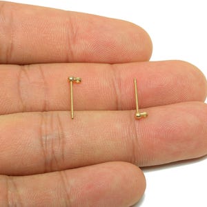 Könnte beinhalten: Zwei kleine, goldfarbene Ohrstecker. Jeder Ohrring hat einen Stecker und ein kleines, rundes Design. Die Ohrringe werden vor einem neutralen Hintergrund gezeigt.