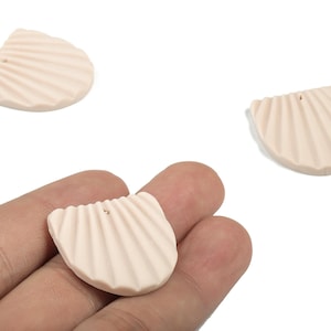 Peut inclure: Trois pendentifs en forme de coquillage de couleur pêche clair avec une texture striée. Un pendentif est tenu dans une main, tandis que les deux autres sont sur fond blanc. Chaque pendentif a un petit trou en haut pour l'enfilage.