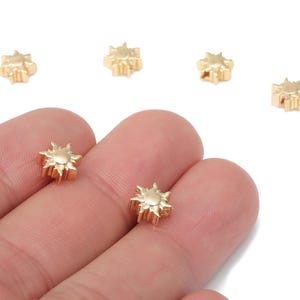 Könnte beinhalten: Nahaufnahme von mehreren kleinen, goldfarbenen sonnenförmigen Perlen. Die Perlen werden in einer Hand vor weißem Hintergrund gehalten. Die sonnenförmigen Perlen haben ein detailliertes Design mit nach außen gerichteten Strahlen.