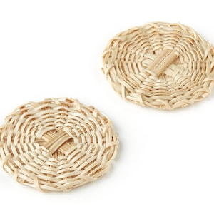 Puede incluir: Dos posavasos redondos de paja trenzada de color beige natural. Los posavasos tienen un patrón tejido con un centro elevado.