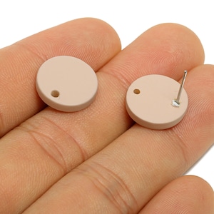 May include: Two round, light beige stud earrings. One earring has a post attached to the back. The earrings are shown on a person's hand. The earrings have a small hole near the edge.