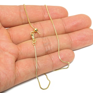 Op de afbeelding: Een goudkleurige boxketting met een verstelbare sluiting. De ketting wordt in een hand gehouden tegen een witte achtergrond. De ketting is een delicaat, fijn sieraad.