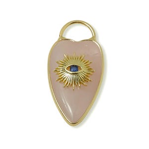 May include: A heart-shaped pendant with a gold-colored frame and bail. The pendant features a light pink stone with a gold sunburst design and a blue eye in the center. The pendant is approximately 2 inches long.