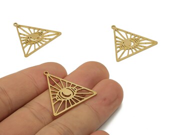 Peut inclure: Charm triangulaire en métal doré avec un motif d'œil. Les breloques sont découpées avec un motif de rayons de soleil autour de l'œil. Une breloque est tenue dans une main, avec deux autres visibles en arrière-plan.