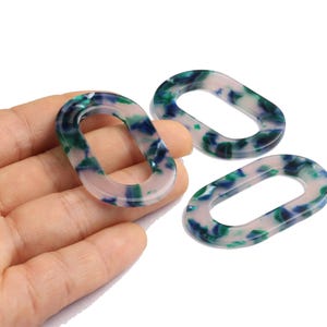 Peut inclure: Trois maillons ovales en acrylique avec un motif marbré bleu, vert et blanc. Un maillon est tenu dans une main, tandis que deux autres sont disposés à proximité sur une surface blanche. Ces maillons pourraient être utilisés pour la bijouterie.