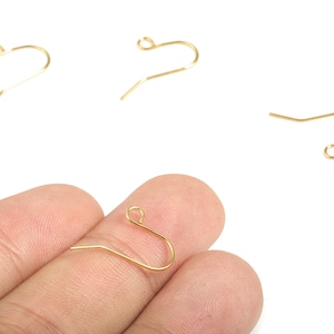 Op de afbeelding: Close-up van verschillende goudkleurige oorbelhaken. Eén haak wordt tussen de vingers gehouden, terwijl drie andere op een witte achtergrond verspreid liggen. De haken hebben een eenvoudig, gebogen ontwerp, ideaal voor het maken van sieraden.