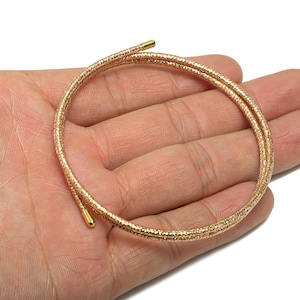 May include: A gold-coloured, textured necklace or bracelet, coiled into a circular shape. The jewellery has a textured surface and is held in a person's hand against a white background. The ends of the jewellery have small, gold-coloured caps.