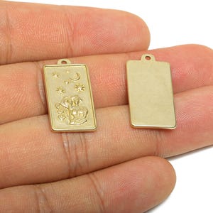 May include: Two gold-colored rectangular charms. One charm features an embossed design of a moon, stars, and two figures. The other charm is plain. Both charms have a small hole at the top for stringing.