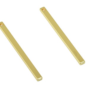 Puede incluir: Dos colgantes de barra de metal dorado con un agujero en la parte superior para sujetar a una cadena o cordón.