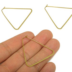 May include: Gold-colored, geometric triangle hoop earrings. One earring is held in a hand, while two others are displayed separately. The earrings have a simple, minimalist design, with a triangular shape and a hook closure. The earrings are made of a shiny, metallic material.