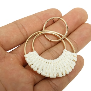 May include: Gold-tone metal earrings with a woven white design. The earrings feature a circular design with a smaller circle attached at the top. The woven section is a half-circle shape. The earrings are held in a hand.