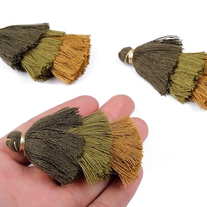 Puede incluir: Borlas de tres niveles en tonos verde oliva y dorado, con una tapa dorada. Las borlas se muestran sobre un fondo blanco, con una de ellas sostenida en una mano. Estas borlas decorativas son adecuadas para diversos proyectos de manualidades.