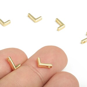 May include: Gold-colored, L-shaped jewelry findings. Several pieces are scattered on a white background, with two held between fingers. Each finding has a small hole for attaching to other components.