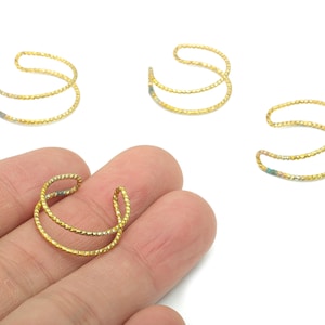 May include: Gold-colored ear cuffs with a double-band design. The ear cuffs have a textured, twisted rope appearance. One cuff is held in a hand, with three others scattered on a white surface. Jewelry.