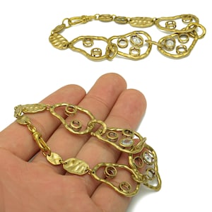 May include: Gold-toned metal bracelet with an abstract design. The bracelet features irregular, openwork shapes with small, clear stones and hammered oval links. One bracelet is displayed on a white background, and another is held in a hand.