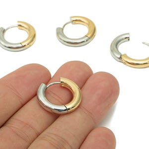 May include: Close-up of several two-tone hoop earrings. Each earring combines gold and silver, with a hinged closure. One earring is held in a hand, showcasing the design. The earrings are on a white background.
