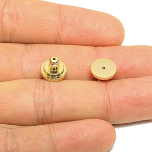Peut inclure: Deux clous d'oreilles et leurs fermoirs dorés sont présentés. Les clous ont un design à gradins avec un petit cylindre surélevé au sommet. Les fermoirs sont ronds et plats. Les articles sont présentés sur un fond neutre.