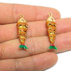May include: Two gold-colored fish-shaped pendants with orange and yellow scales, red accents, and green and red tail fins. Each pendant has a loop at the top for hanging. The pendants are displayed against a light background.