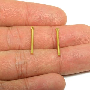 Può includere: Due piccoli ciondoli a forma di barra in metallo color oro. Ogni ciondolo è una sottile barra rettangolare con un piccolo anello nella parte superiore. I ciondoli sono tenuti nella mano di una persona su sfondo bianco.