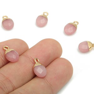 Op de afbeelding: Roze edelsteenbedels met goudkleurige doppen en lussen. De bedels zijn ovaal van vorm en hebben een facetgeslepen oppervlak. Verschillende bedels zijn verspreid op een witte achtergrond, sommige worden in een hand gehouden.